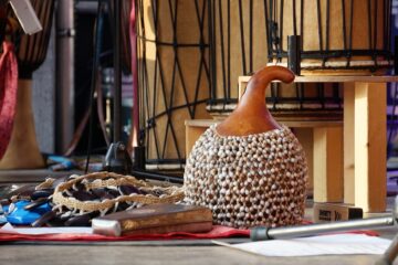 Shekere und verschiedene Kleinpercussion-Instrumente liegen auf dem Boden, im Hintergrund sind Basstrommeln zu sehen.
